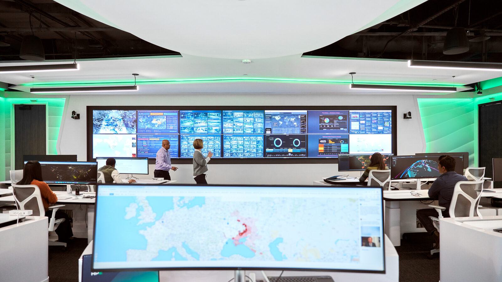 A group of people is engaged in a discussion in a modern control room with multiple large screens displaying data and maps.